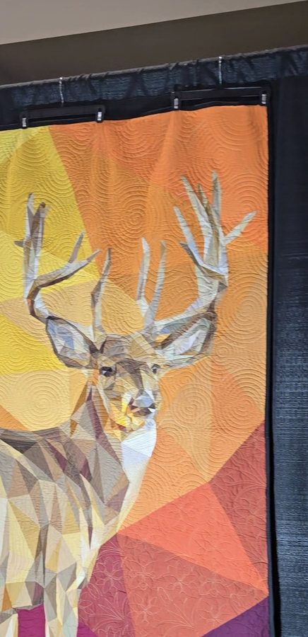 A paper-pieced stag quilt in greys and creams against a sunset gradient of orange, yellow and pink