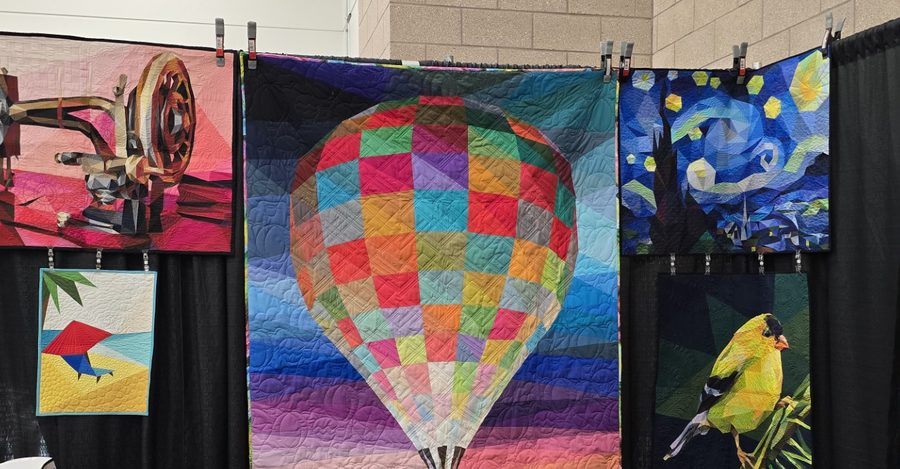 A row of Legit Kits quilts hanging side by side, including a patchwork hot air balloon and a paper-pieced night sky