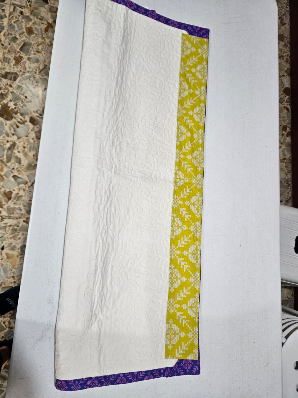 A finished Loter&iacute;a quilt folded on a work table, showing the quilted reverse, the yellow patterned strip and the purple and pink binding