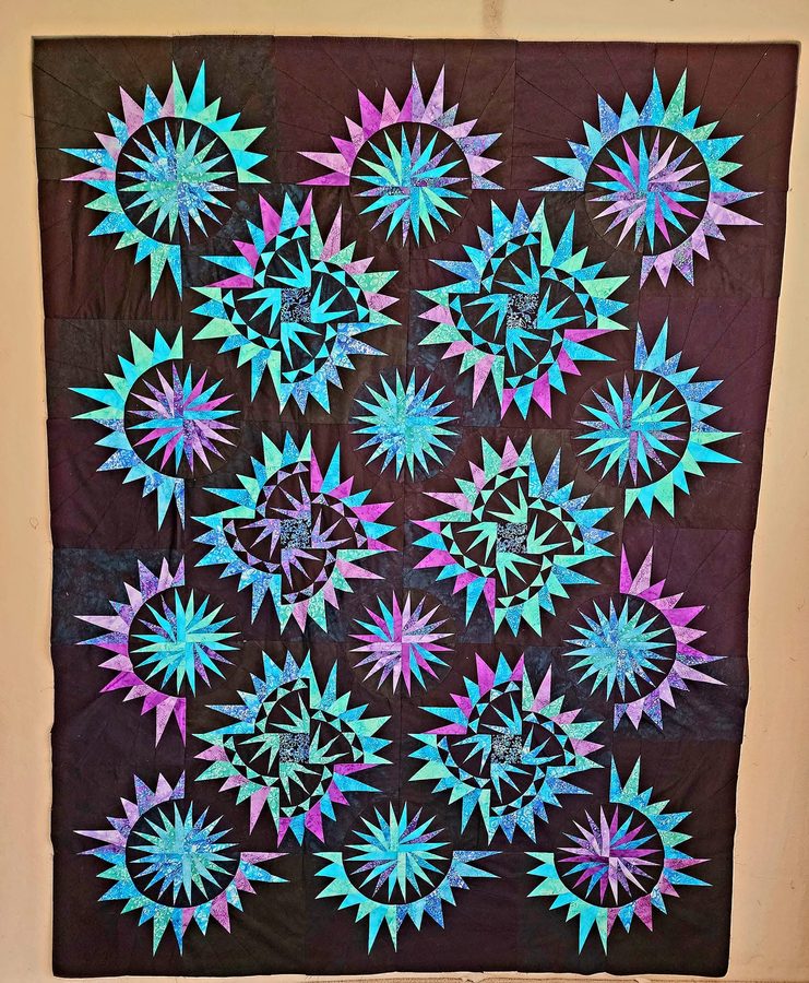 A quilt of turquoise and violet starbursts scattered across a black ground